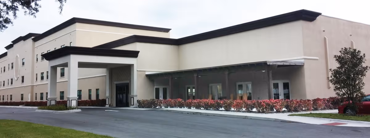 Front entrance and facade of a beige multi-story senior living building with a covered drop-off driveway and landscaped beds.