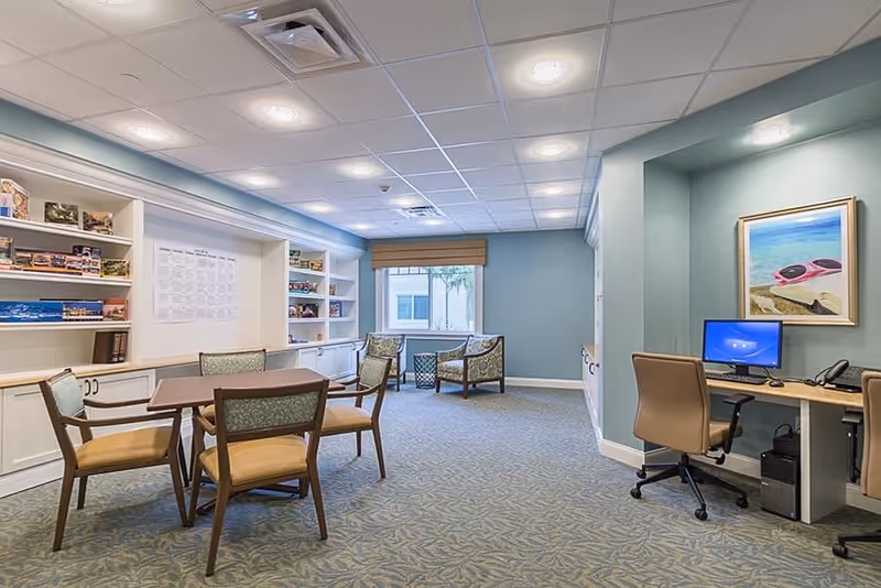 A well-lit room with light blue walls and patterned carpet featuring a table with four chairs in the center, bookshelves filled with games and books on the left wall, two armchairs near a window at the back, and a desk with a computer and office chair on the right side beneath a framed beach-themed painting.