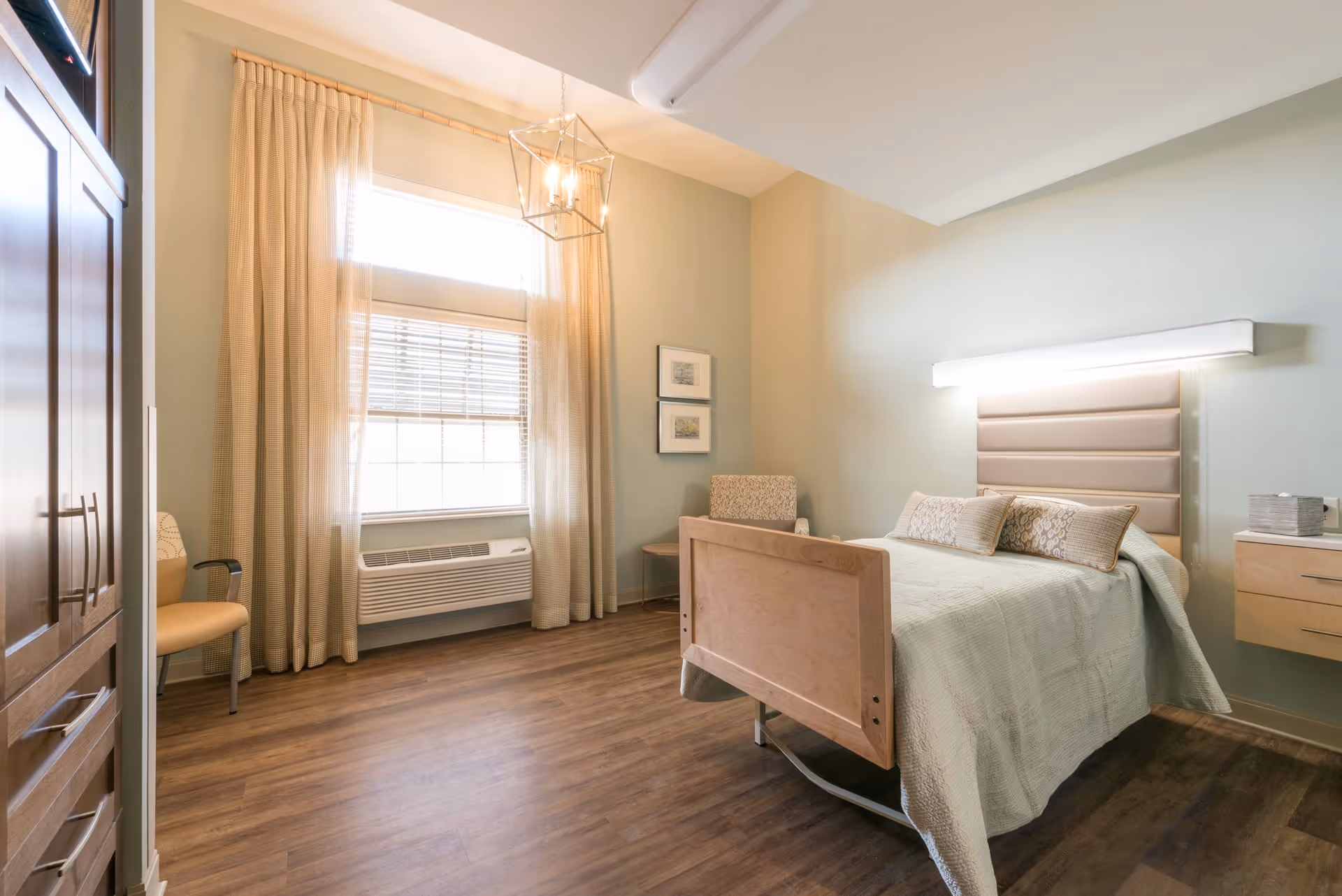 A bright and clean nursing facility bedroom with a single bed featuring a padded headboard and light green bedding. The room has wooden flooring, a large window with beige curtains, a beige armchair, a small side table, and a wooden wardrobe. The walls are painted a soft green color, and there is a modern light fixture hanging from the ceiling.