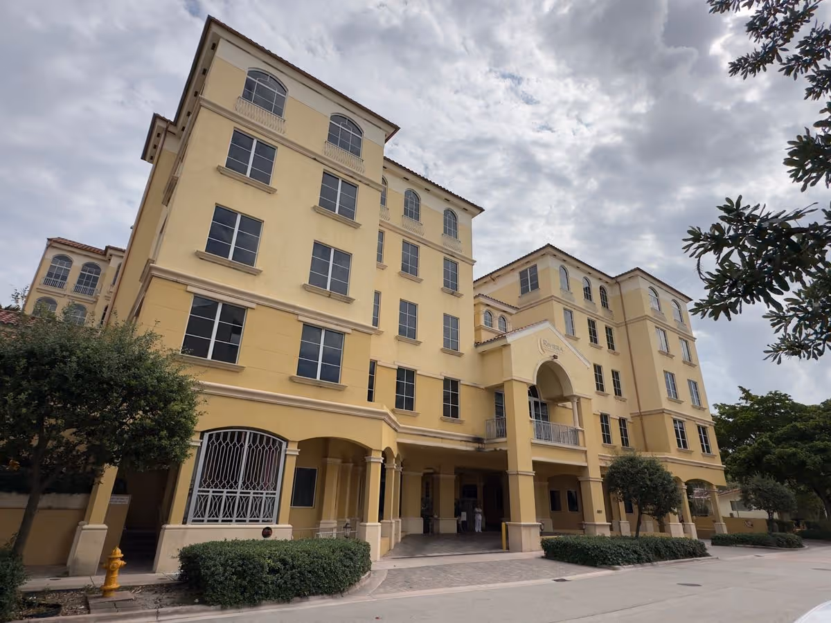 Front exterior of a yellow, multi-story Mediterranean-style building with arched entrance and balconies under a cloudy sky.