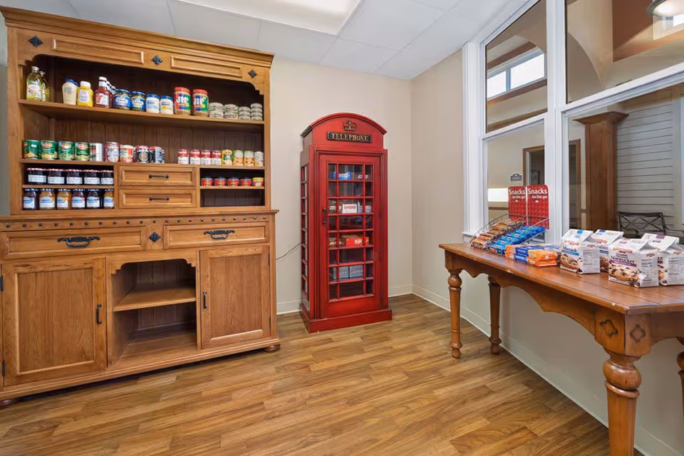 Interior room with wooden flooring featuring a large wooden cabinet stocked with various canned and jarred food items, a red vintage-style telephone booth, and a wooden table displaying assorted packaged snacks near a window with white trim.