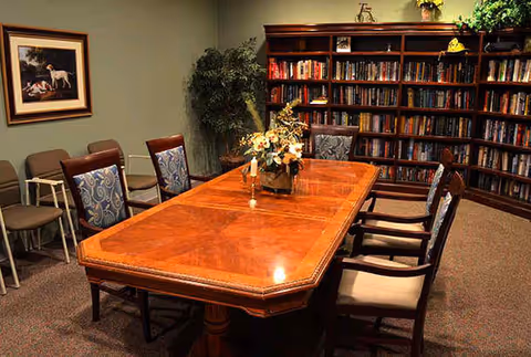 A cozy room with a large wooden table surrounded by chairs, a bookshelf filled with books along the back wall, a framed painting on the left wall, and a potted plant in the corner.