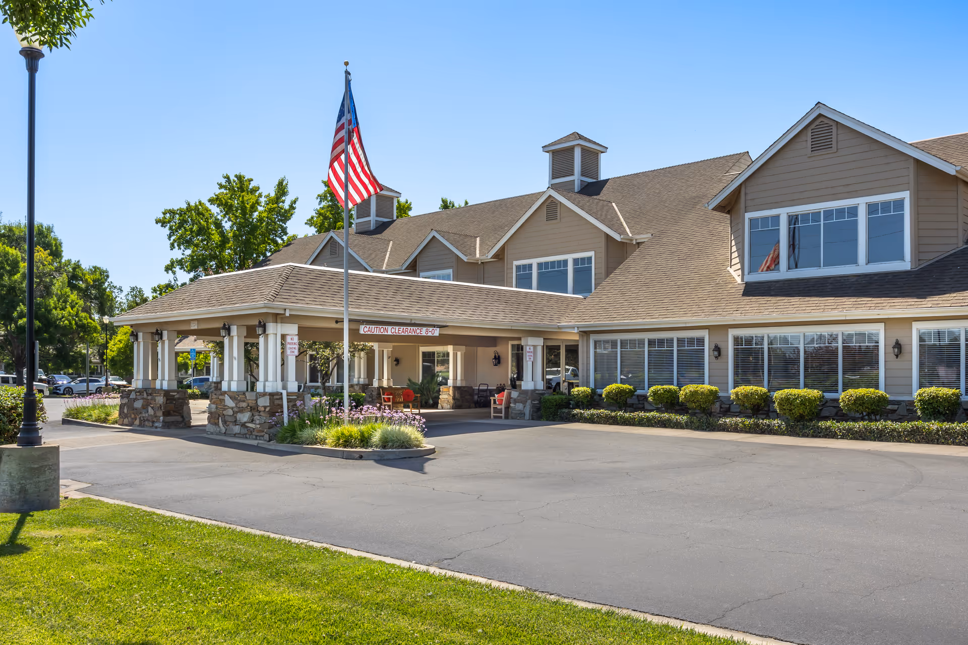 Exterior view of Brookdale Lodi senior living facility showing the entrance with a covered drop-off area, an American flag on a flagpole, well-maintained landscaping, and a clear blue sky.