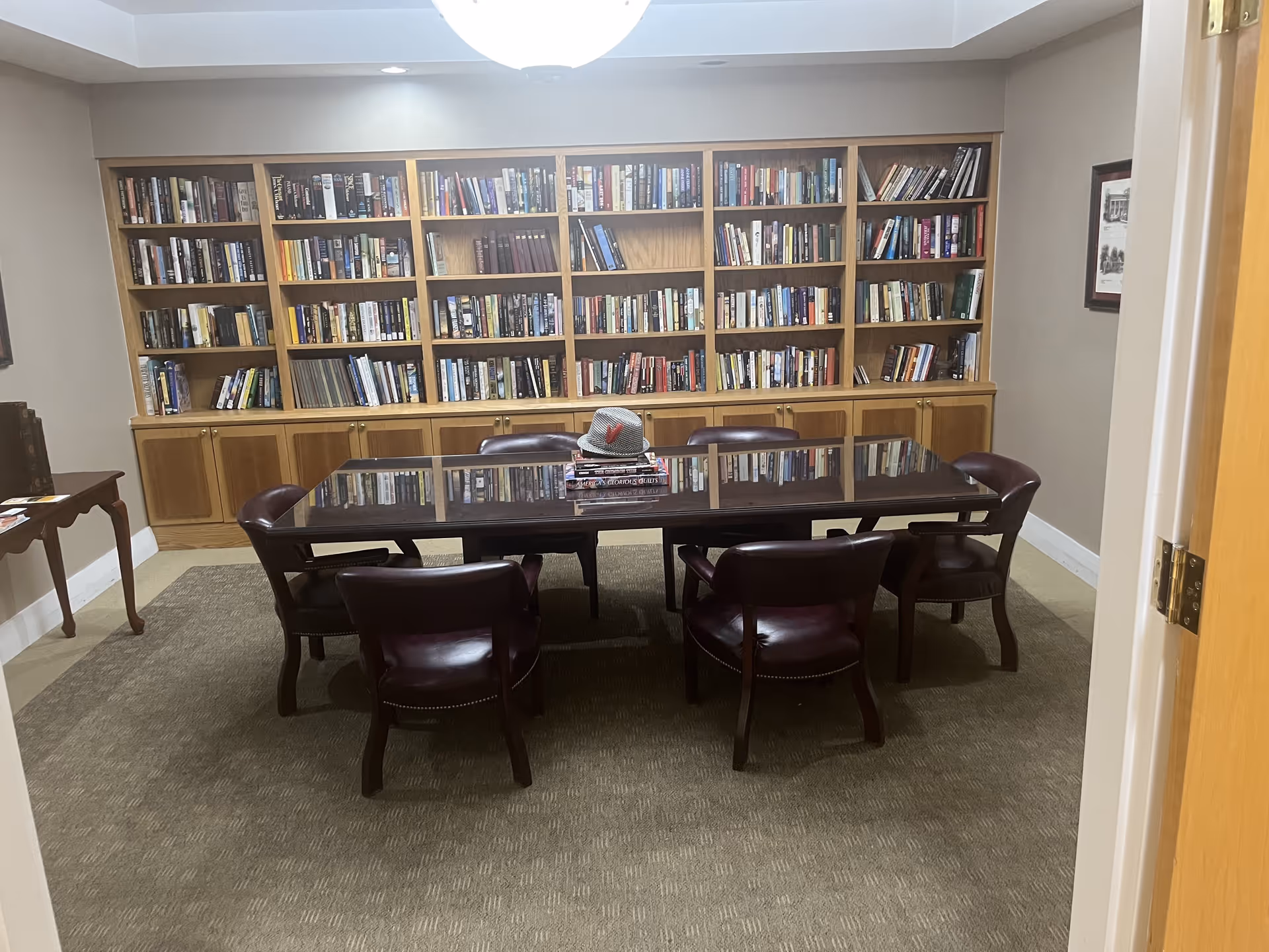 A quiet library room with a large wooden table surrounded by six dark leather chairs. Behind the table is a built-in wooden bookshelf filled with books. A hat and a few stacked books are placed on the table. The room has beige walls and carpeted floor.
