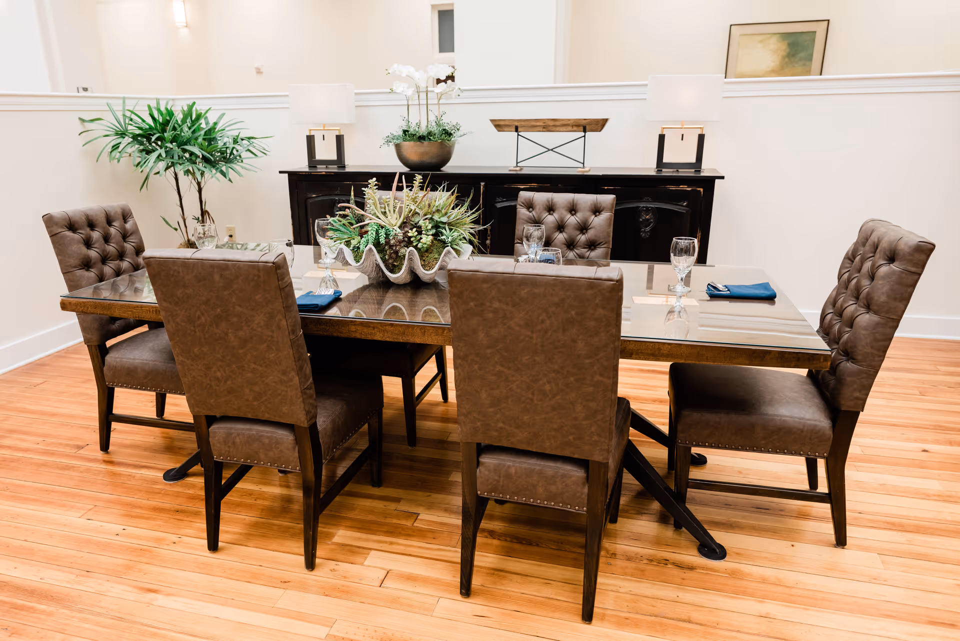 A dining room with a rectangular glass-top table surrounded by six brown upholstered chairs. The table is set with glassware and blue napkins. A large decorative centerpiece with succulents is in the middle of the table. In the background, there is a black sideboard with two table lamps, a potted plant, and a framed picture on the wall. The room has light-colored walls and wooden flooring.