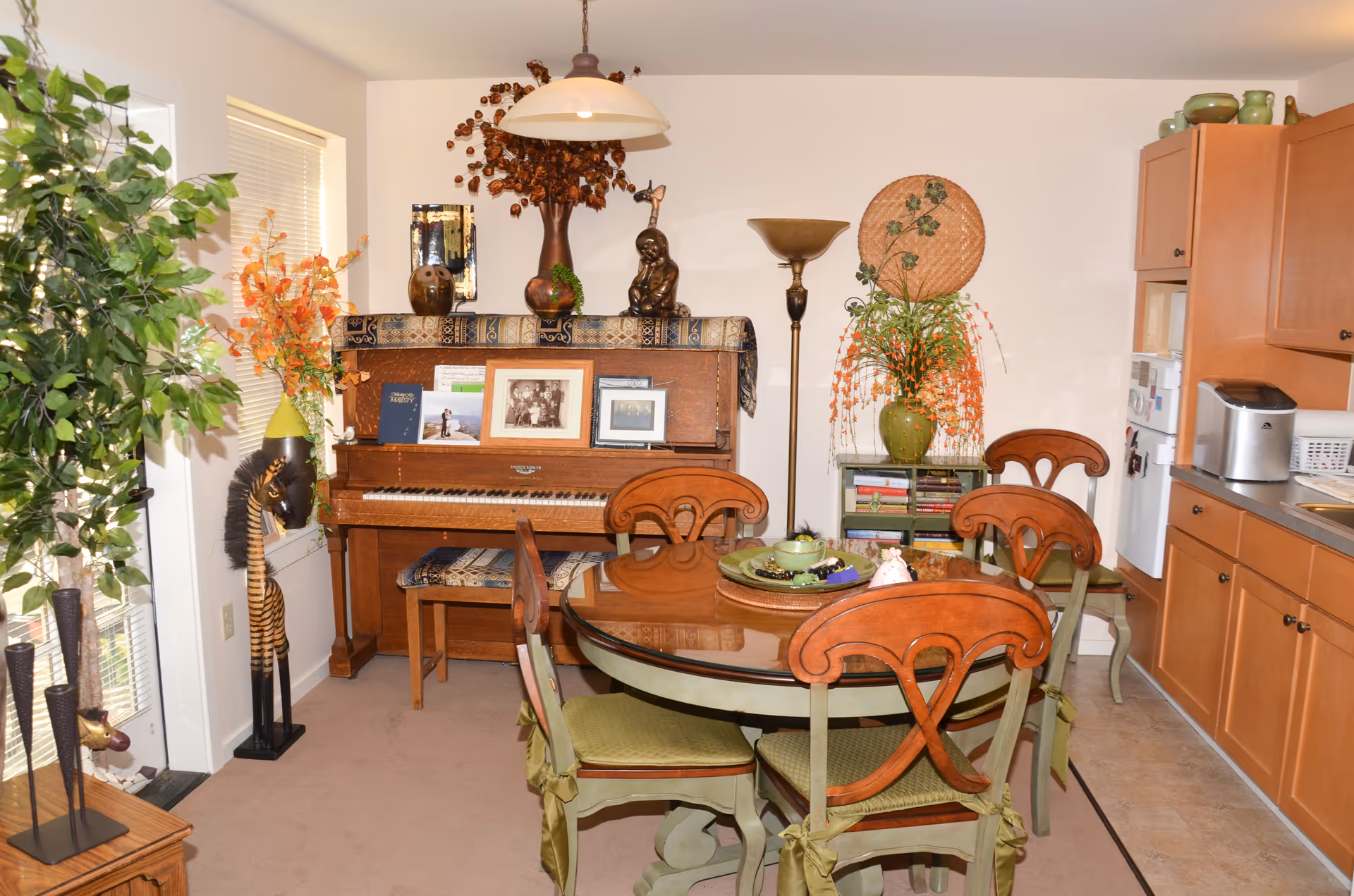 A cozy dining area with a round glass-top table surrounded by four wooden chairs with green cushions. Behind the table is an upright piano decorated with framed photos, books, and ornamental vases. To the right, there is a small bookshelf with a large vase holding orange flowers and a woven decorative piece on the wall above it. The kitchen area with wooden cabinets, a refrigerator, and a countertop with a toaster is visible on the right side. On the left, there is a tall green plant and a decorative giraffe sculpture near a window with blinds.