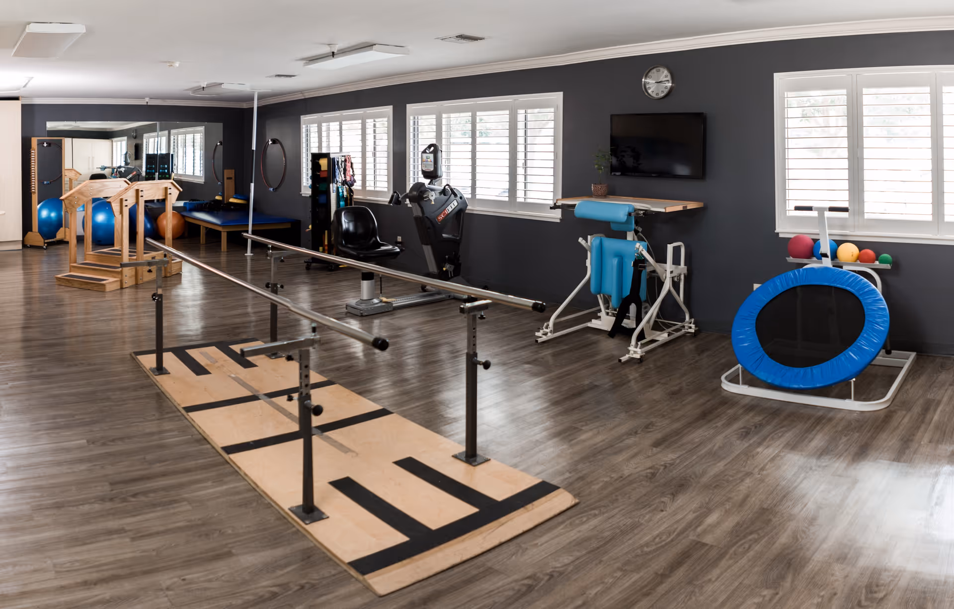 A spacious physical therapy room with wooden flooring, parallel bars for walking exercises, exercise balls, a small trampoline, various therapy equipment, a wall-mounted TV, and large windows with white shutters letting in natural light.