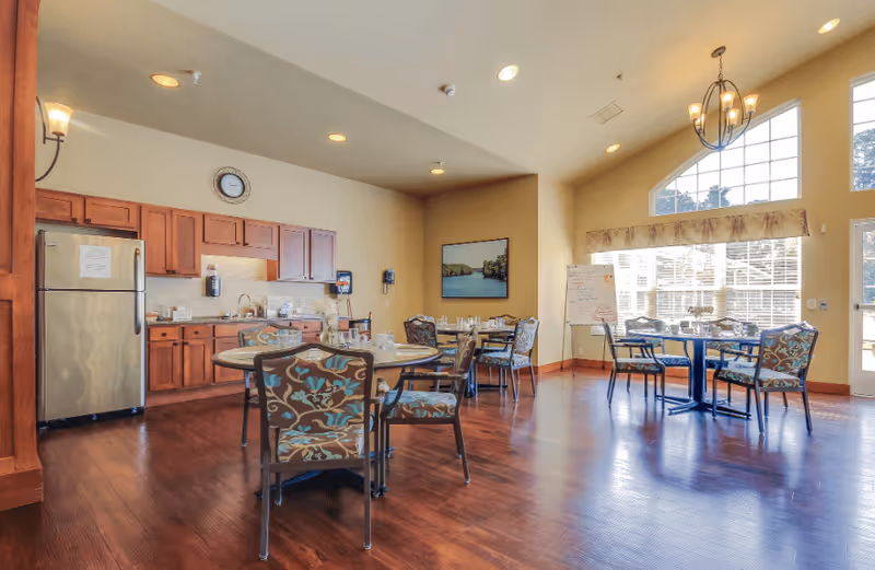 A bright dining area in an assisted living facility featuring several round tables with floral upholstered chairs. The room has wooden flooring, a kitchen area with wooden cabinets and a stainless steel refrigerator, large windows letting in natural light, a chandelier, and a whiteboard in the corner.