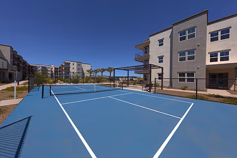 A blue outdoor tennis/pickleball court in front of multi-story apartment buildings under a clear blue sky.
