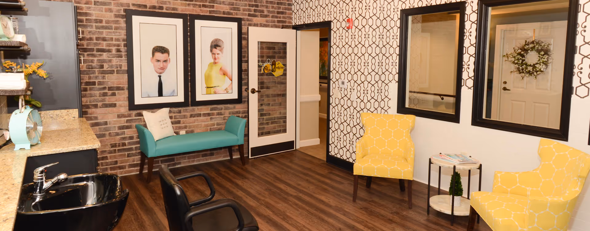 A cozy interior room with wood flooring and a mix of brick and patterned wallpaper walls. The room features a turquoise bench with a pillow, two yellow patterned armchairs, a small round table with magazines and a small plant, and a black salon-style chair near a black sink. Two large framed portraits hang on the brick wall, and two large mirrors are mounted on the patterned wallpaper wall. A door with a glass panel and a decorative sign is visible in the background.