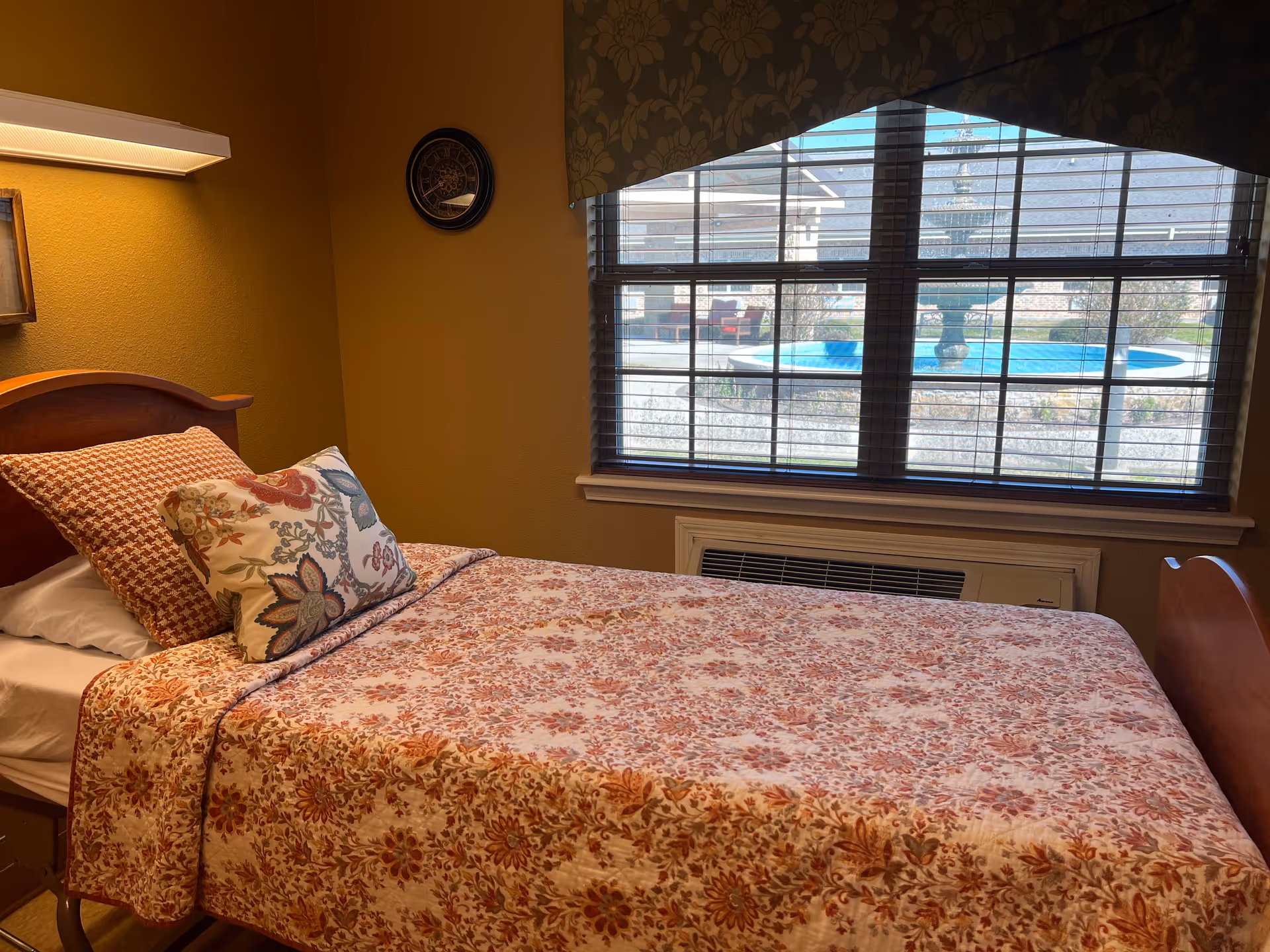 Single bed with floral bedding and decorative pillows beside a large window overlooking an outdoor fountain area.