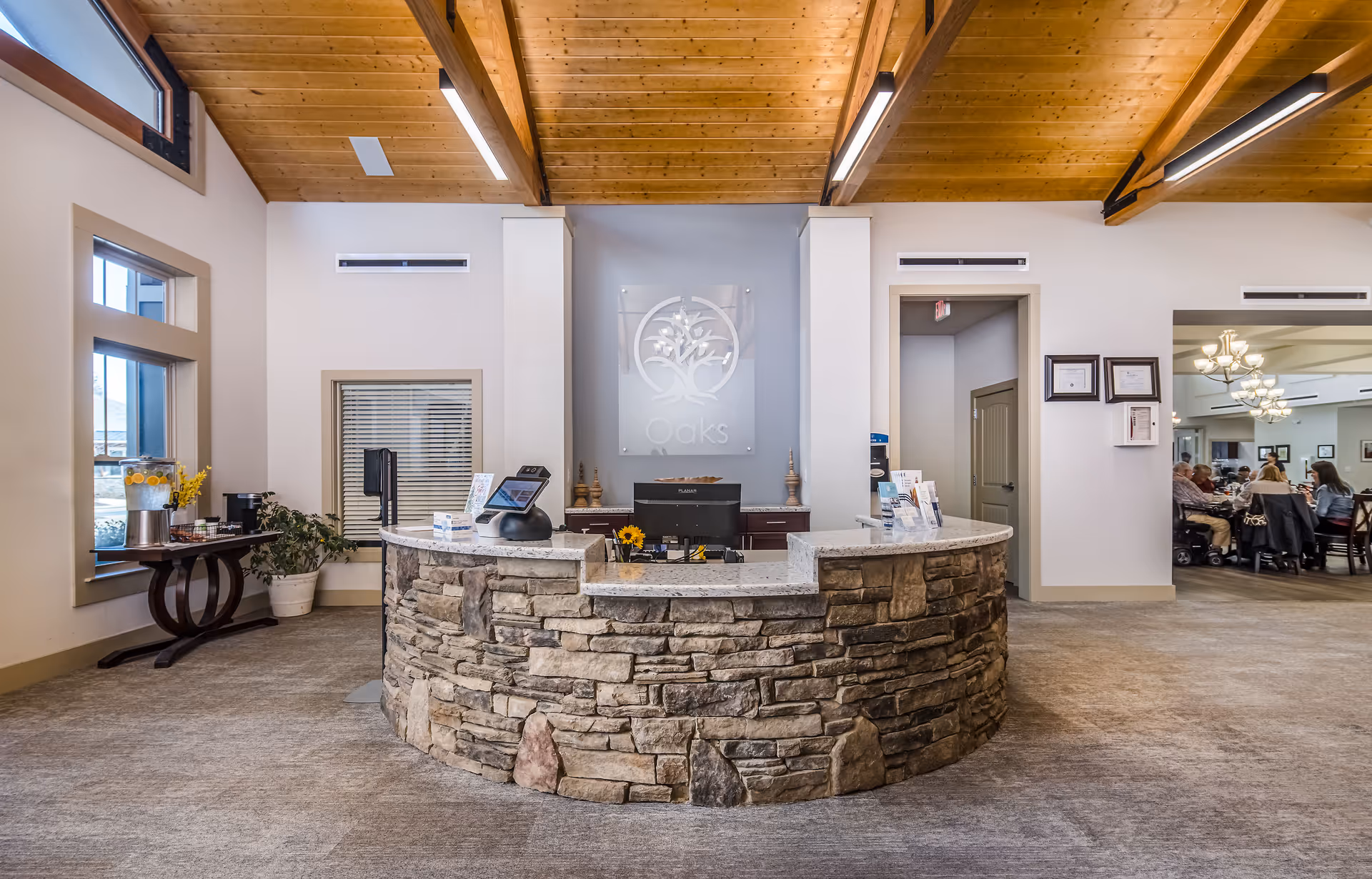 Stone-front reception desk in a bright lobby with wood-beamed ceiling and a dining area visible to the right.