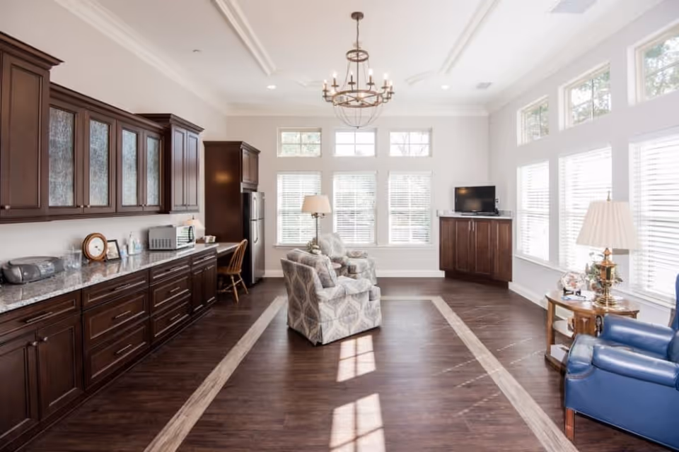 Bright and spacious common area with large windows letting in natural light. The room features dark wood cabinetry along one wall with a granite countertop, a small desk with a chair, and a stainless steel refrigerator. In the center, there are two patterned armchairs facing a corner cabinet with a flat-screen TV. A blue armchair and a side table with a lamp are positioned near the windows. The floor is dark wood with a lighter wood border, and a chandelier hangs from the ceiling.