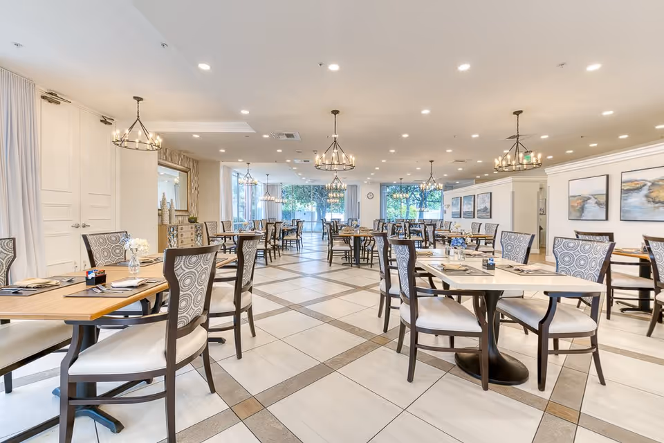 A spacious, well-lit dining room with multiple tables and chairs arranged neatly. The room features large windows allowing natural light to enter, elegant chandeliers hanging from the ceiling, and decorative artwork on the walls. Tables are set with placemats, napkins, and small flower arrangements.