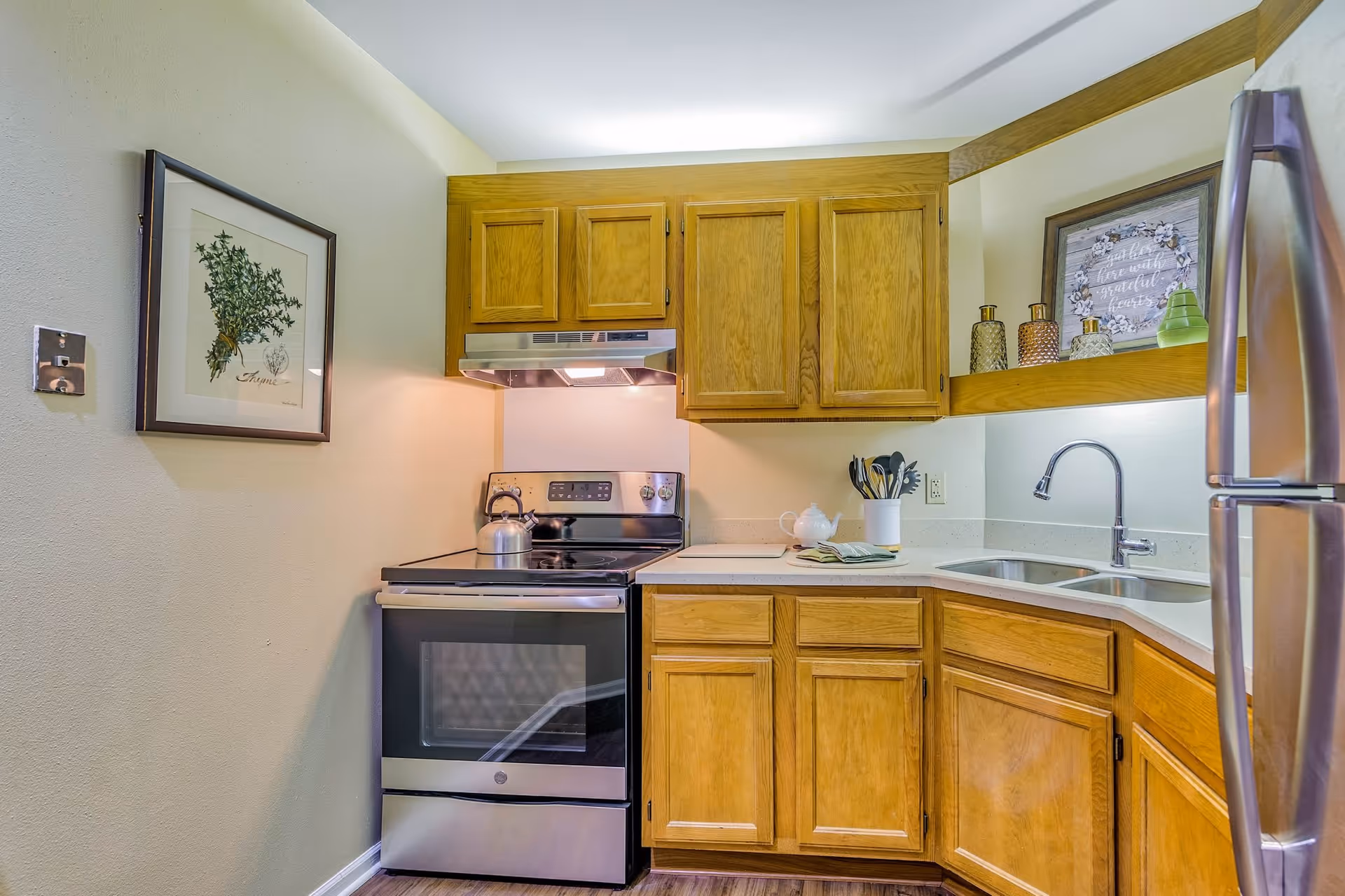 A compact kitchen with wooden cabinets, a stainless steel electric stove with a kettle on top, a double sink with a modern faucet, and a stainless steel refrigerator. The kitchen has a light-colored countertop and a framed botanical print on the wall. Decorative bottles and a framed sign are placed on a shelf above the sink.