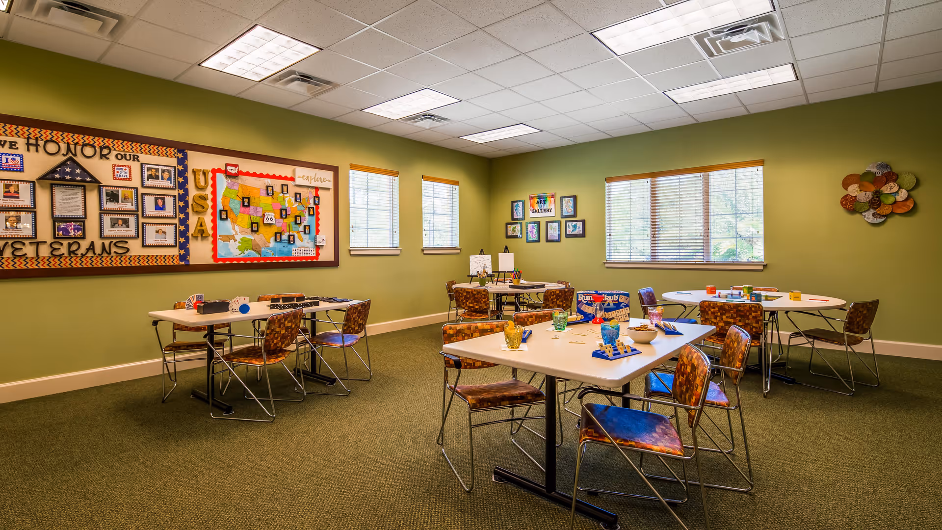 A brightly lit activity room with green walls and carpeted floor featuring several tables and chairs arranged for group activities. The tables have board games and colorful cups on them. The walls display a large bulletin board honoring veterans, a map of the USA, and framed artwork. Three windows with blinds allow natural light into the room.