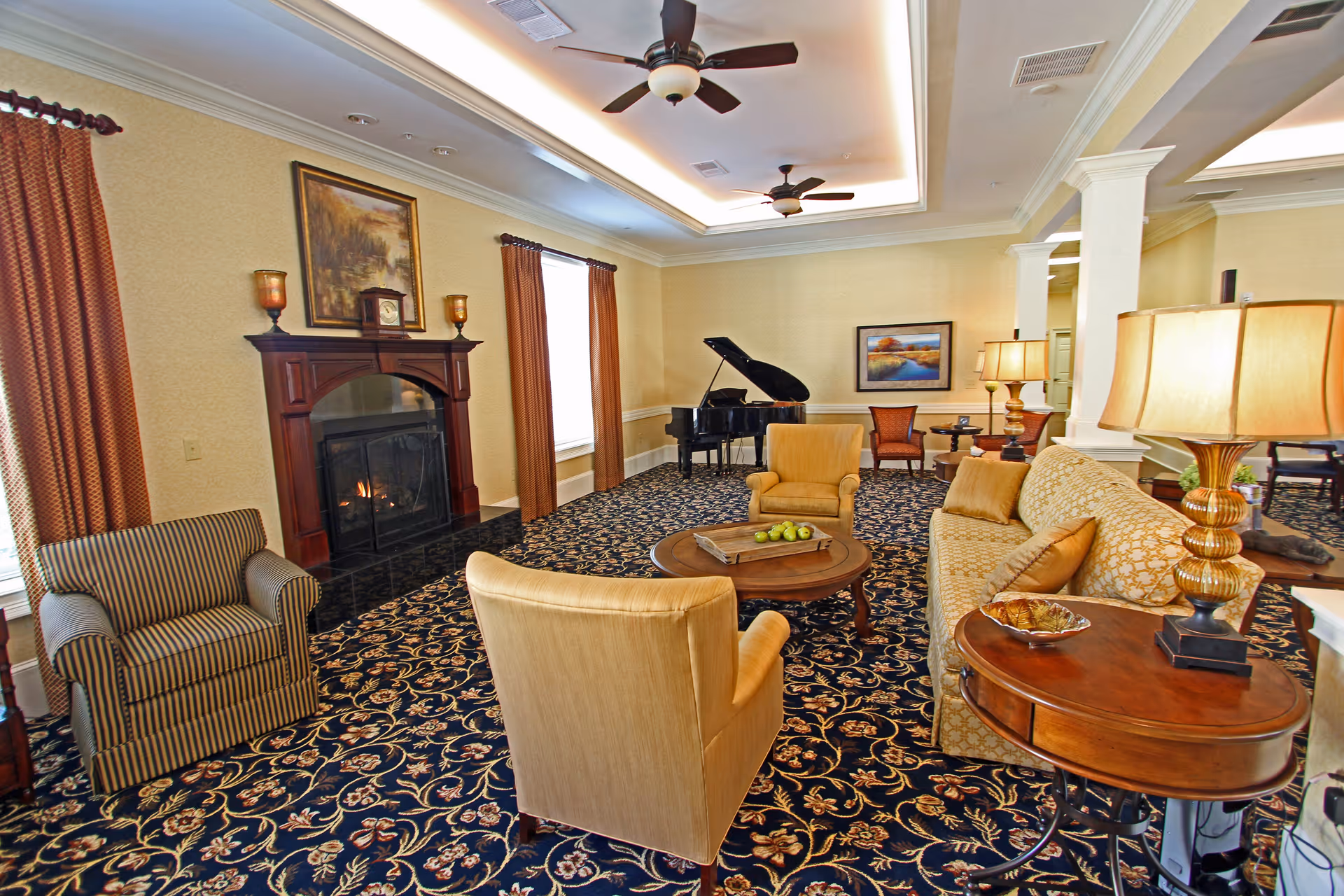 A cozy living room with patterned carpet, a fireplace with a wooden mantel, two large windows with brown curtains, a grand piano in the corner, and comfortable seating including a striped armchair, two beige armchairs, and a patterned sofa. There are two table lamps on wooden side tables and framed artwork on the walls.