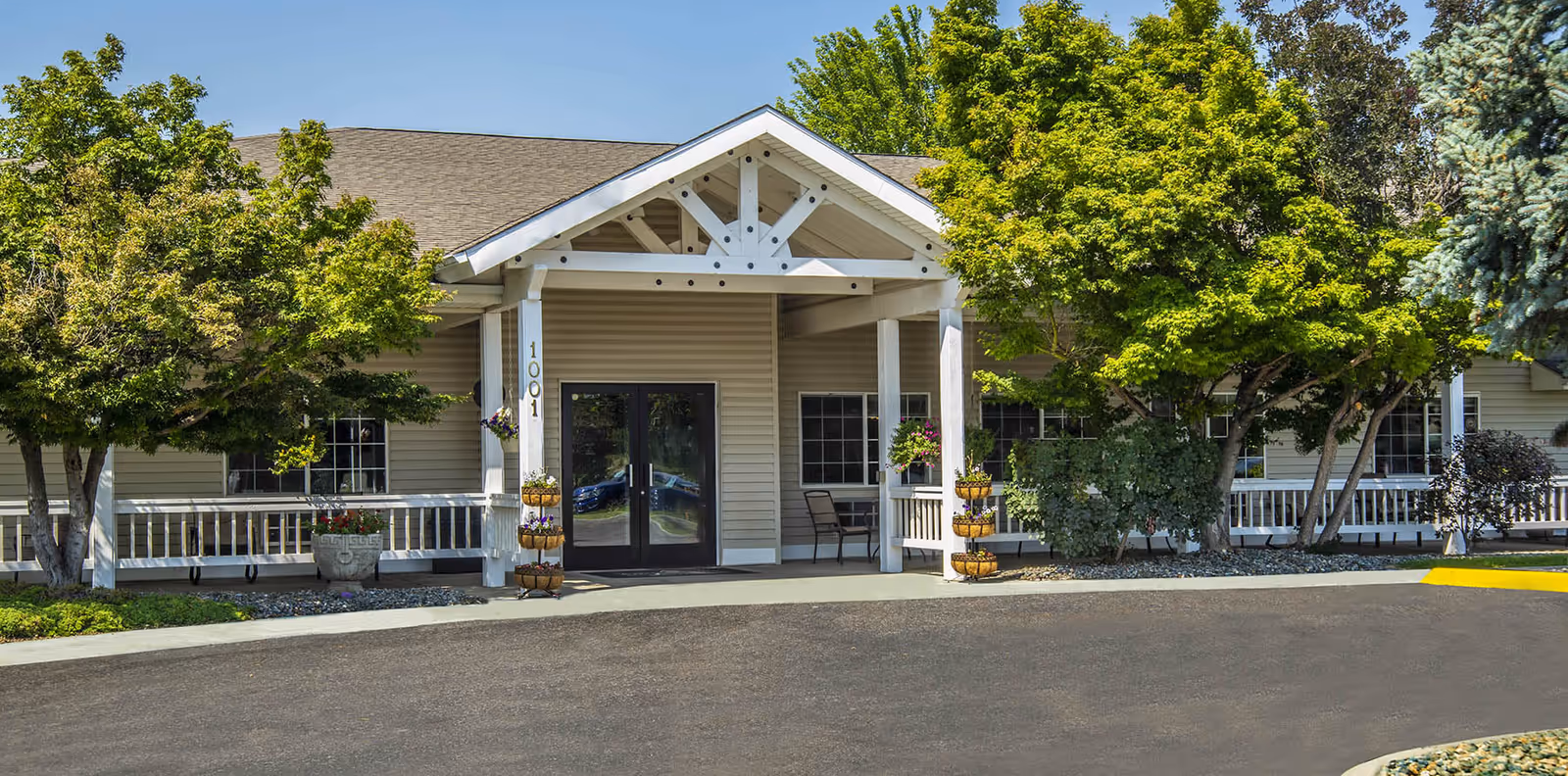 Front exterior view of a single-story senior living facility building with beige siding, a peaked roof, and a covered entrance supported by white columns. The entrance has double glass doors and the number 1001 is displayed on one of the columns. There are trees and shrubs on either side of the entrance, along with a paved driveway in front.