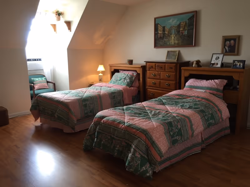 A bedroom with two twin beds covered in matching pink and green patterned bedspreads. Between the beds is a wooden dresser with framed pictures and a small lamp on it. There is a window with sheer white curtains letting in natural light, and a green cushioned chair is placed near the window. The room has wooden flooring and light-colored walls.