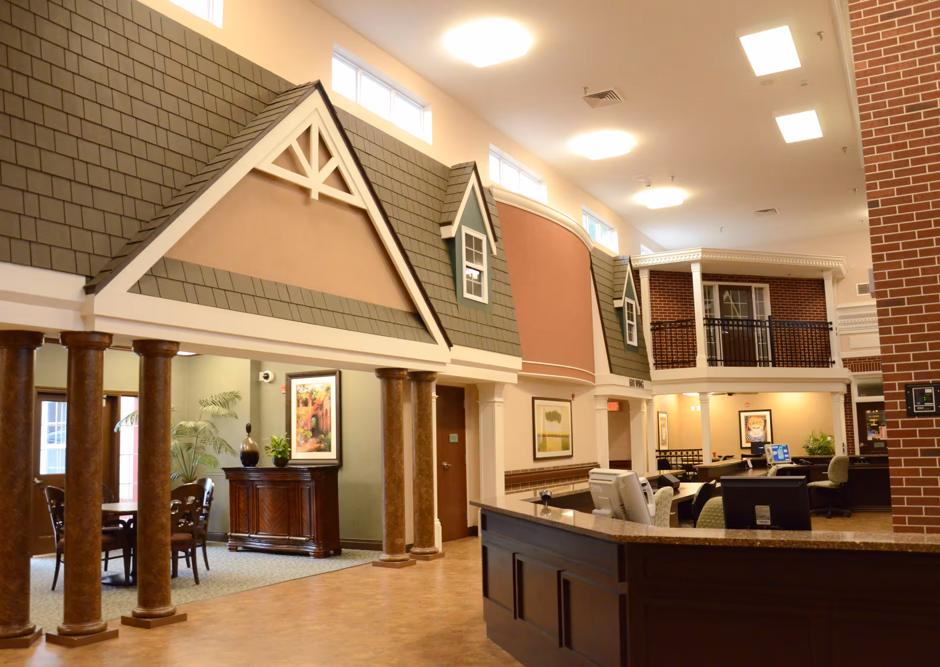 Interior view of Pavilion of Ottawa featuring a reception desk in the foreground, decorative columns, and a hallway with seating areas and framed artwork. The design includes architectural elements resembling house facades with windows and gabled roofs inside the building.