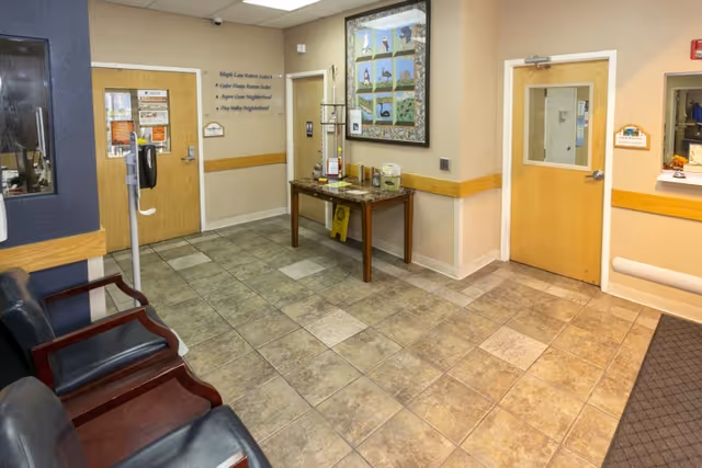 Interior view of a waiting area with tiled floor, beige walls, and wooden trim. There are three black chairs with wooden armrests on the left side, a wooden table with informational brochures and a hand sanitizer dispenser against the far wall, and two wooden doors, one with a window and the other with signage. A large framed artwork hangs on the wall above the table.