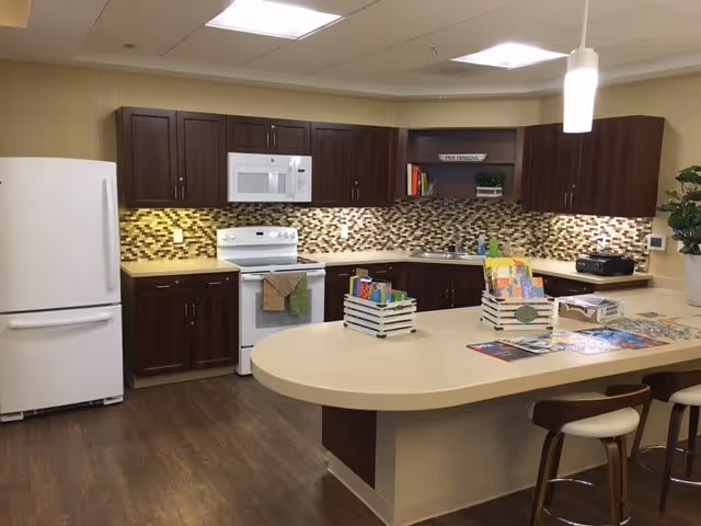 A modern kitchen area in an assisted living facility featuring dark wood cabinets, a white refrigerator, white microwave, and white stove. The backsplash has a mosaic tile design in earth tones. There is a large beige countertop island with stools and various brochures and books displayed on it. The floor is wood, and the lighting is bright with ceiling lights and a hanging pendant light.
