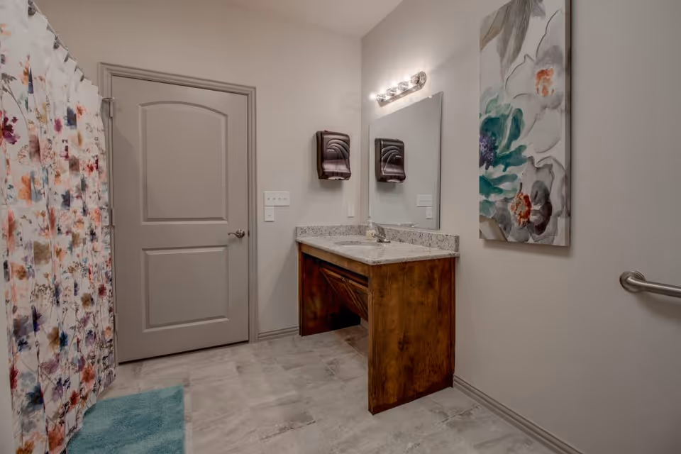 A bathroom with a floral shower curtain on the left, a closed gray door, a wooden vanity with a granite countertop and a sink, a large mirror above the sink, two wall-mounted soap or paper towel dispensers, a colorful floral painting on the wall, and a metal grab bar on the right wall.