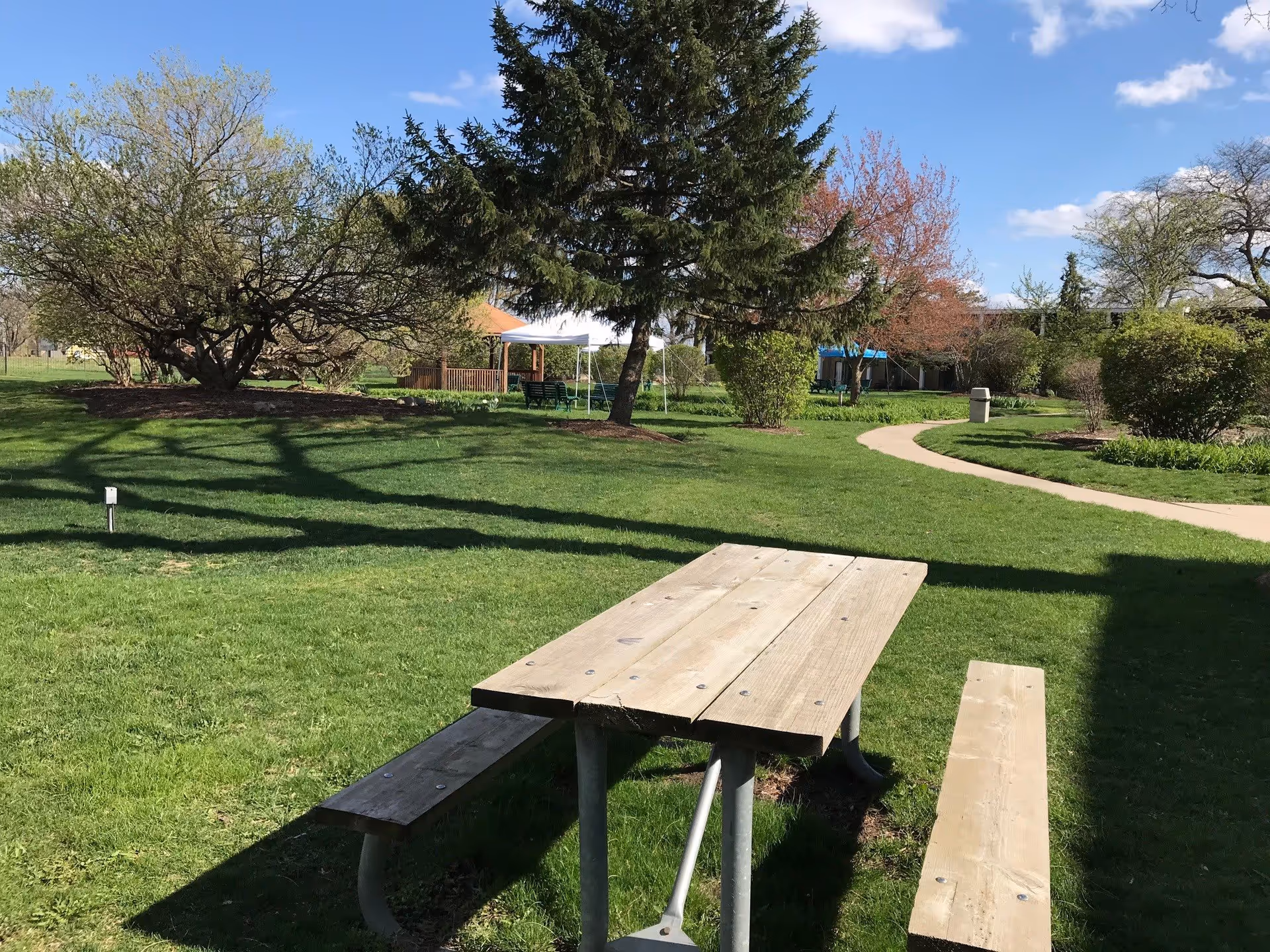 Outdoor scene at Asbury Gardens featuring a wooden picnic table with attached benches on a grassy lawn. Surrounding the area are various trees and bushes, a paved walking path, and a gazebo with a white canopy in the background under a partly cloudy blue sky.