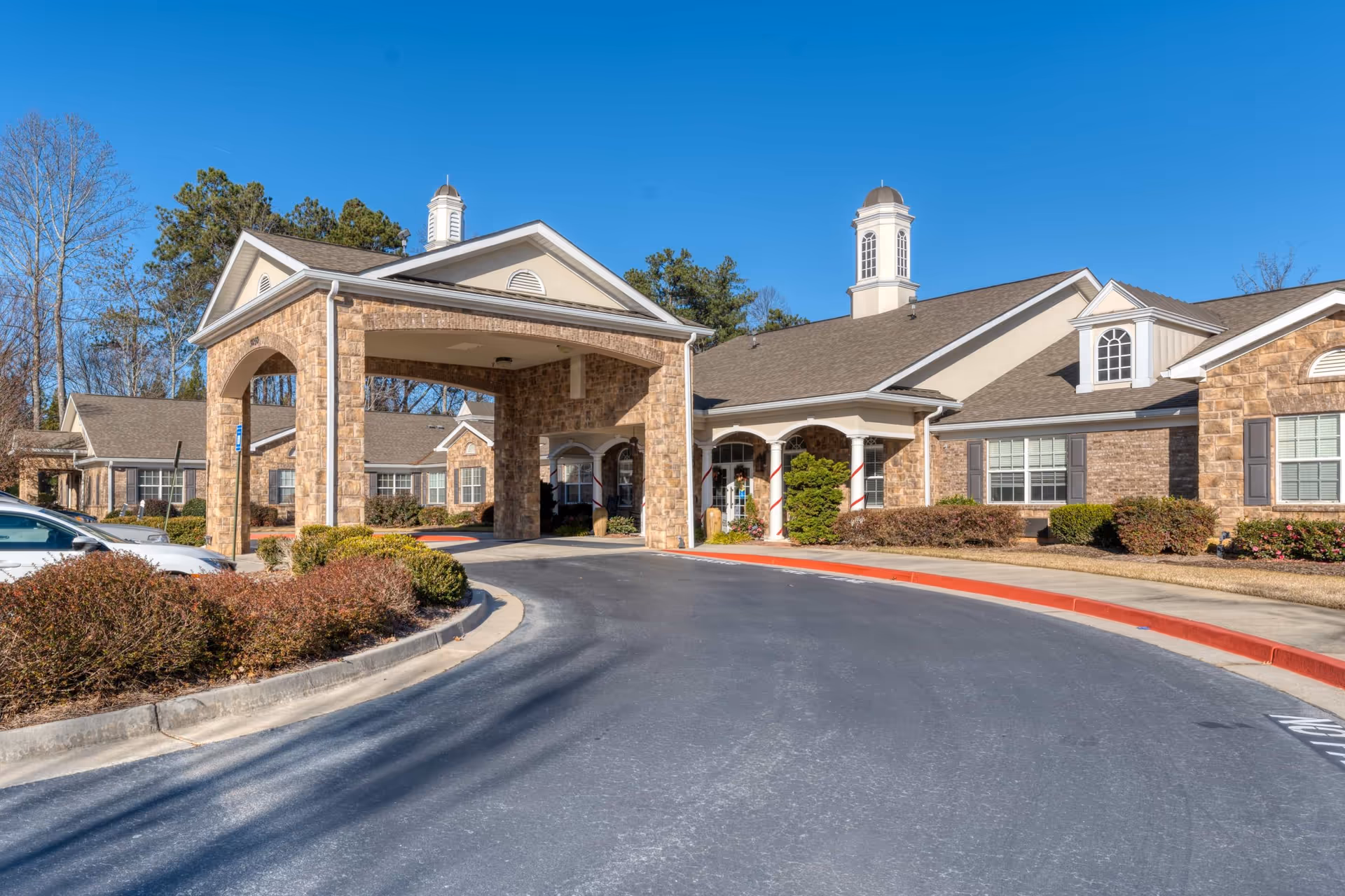 Front exterior view of Addington Place of Johns Creek, showing a covered entrance with stone pillars, a driveway, and landscaped bushes under a clear blue sky.