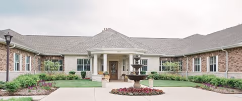 Front exterior view of a single-story brick building with a covered entrance supported by white columns. In front of the entrance is a multi-tiered water fountain surrounded by a circular flower bed. The building is surrounded by well-maintained landscaping with green grass, shrubs, and flowers under a cloudy sky.