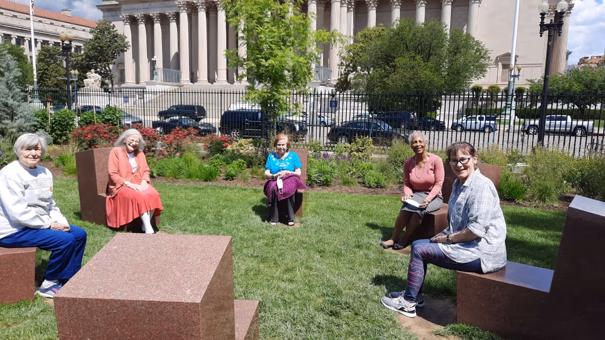 Five elderly women sitting outdoors on stone benches arranged in a circle on a grassy area with bushes and flowers behind them. In the background, there is a large classical building with tall columns and a black iron fence separating the garden from the street with parked cars.