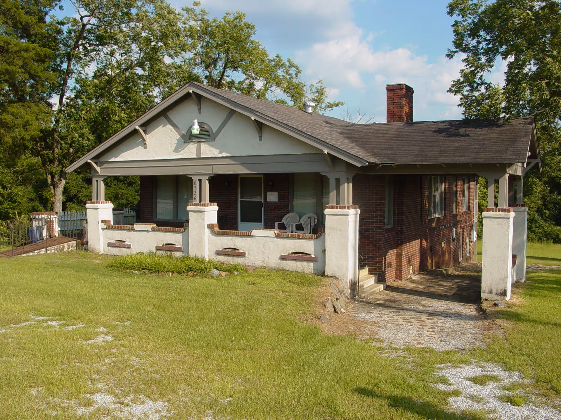 A single-story brick house with a covered front porch featuring white pillars and a small seating area with two chairs. The house is surrounded by green grass and trees under a partly cloudy sky.