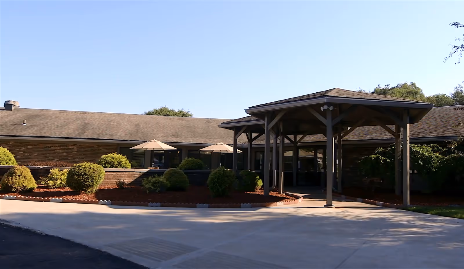 Exterior view of Corry Manor senior living facility showing a single-story brick building with a covered entrance supported by wooden beams, surrounded by landscaped bushes and small trees under a clear blue sky.