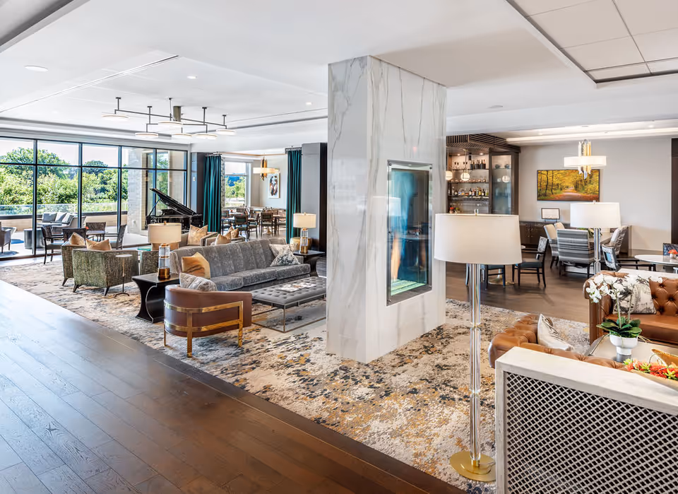 Bright, spacious communal lounge with sofas, armchairs, a central glass-enclosed fireplace, a grand piano, and large windows overlooking trees.