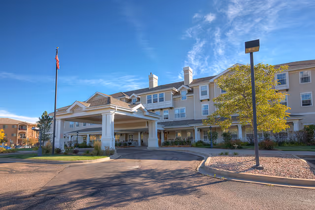 Exterior view of a multi-story senior living facility with a covered entrance driveway, an American flag on a flagpole, a tree, and a clear blue sky.