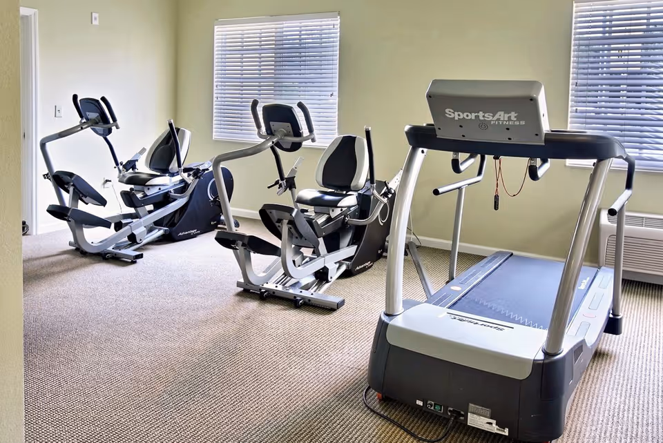 Small fitness room with a treadmill and two recumbent stationary bikes by windows.