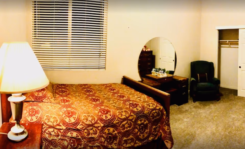 A bedroom with a bed covered in a patterned red and gold bedspread, a wooden nightstand with a lamp, a large round mirror on a wooden dresser, a green armchair, and an open closet with sliding doors. The room has beige walls and carpeted floor.
