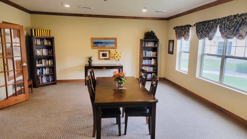 A bright room with a wooden table and six chairs in the center, two bookshelves filled with books against the walls, a small console table with decorative plants and flowers, and three large windows with patterned valances letting in natural light.