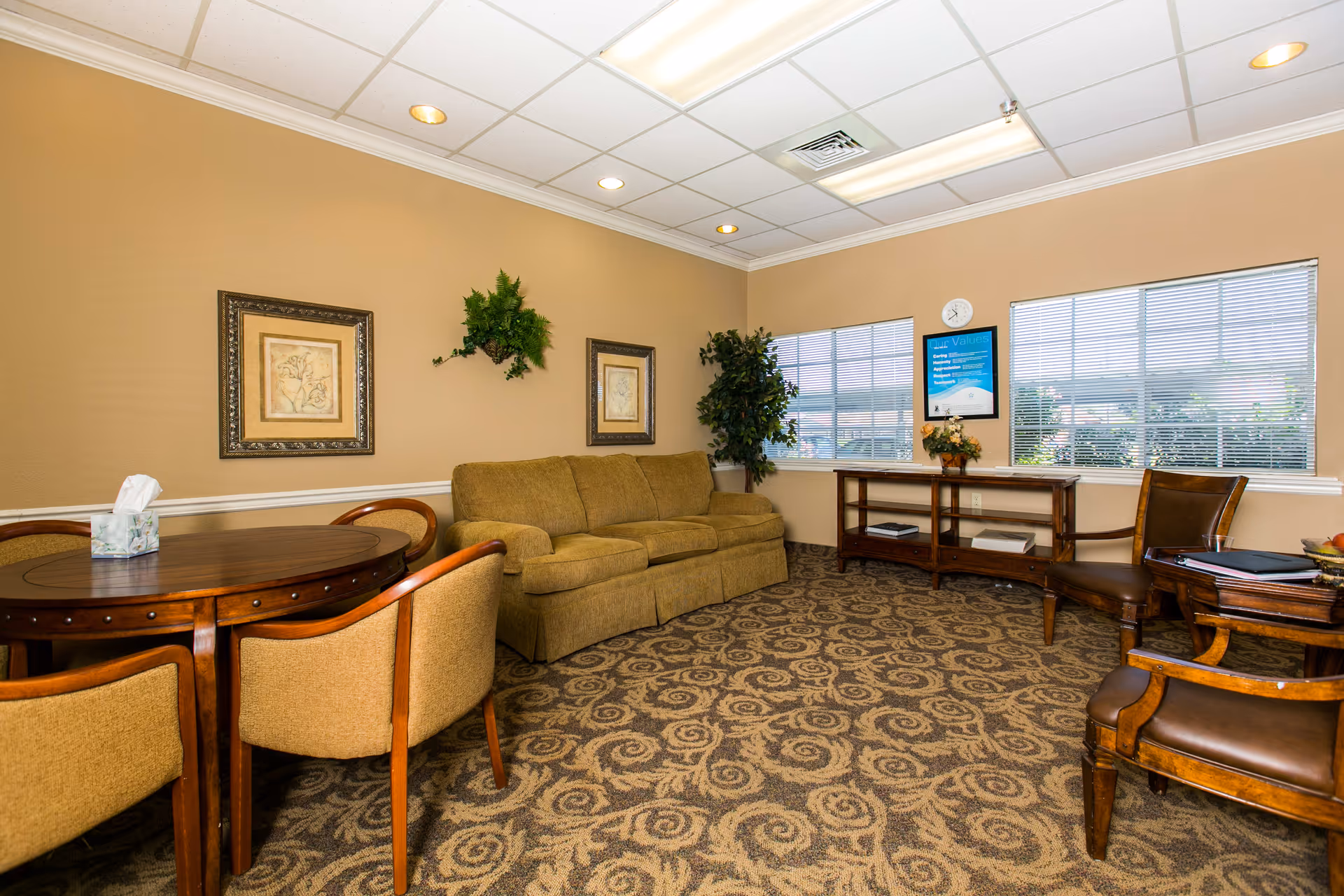 Warmly decorated lounge with a sofa, armchairs, a round table, and windows in a senior living facility.