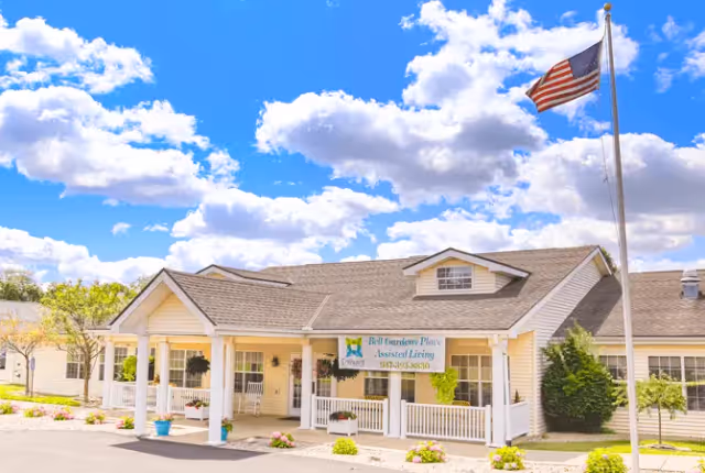 Exterior view of Trustwell Living at Bell Gardens Place, an assisted living facility with a single-story building featuring a covered porch, white railings, and an American flag on a flagpole in front. The sky is blue with scattered clouds.