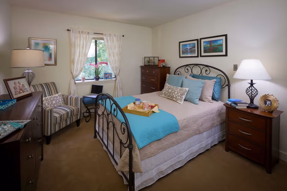 A cozy furnished bedroom featuring an iron-framed bed with teal accents, bedside tables and lamps, a striped armchair, dresser, and a curtained window.