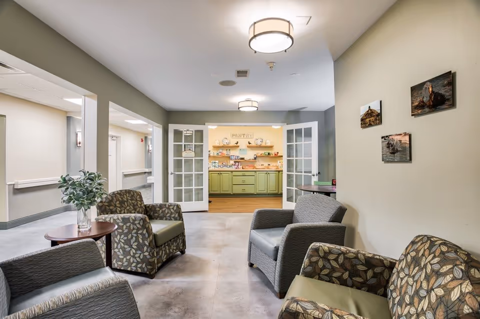 A cozy seating area in a senior living facility with four armchairs arranged around a small round table with a vase of greenery. The walls are light-colored with three framed pictures hanging on one side. In the background, double glass doors open to a pantry area with green cabinets and shelves stocked with snacks and items. The space is well-lit with ceiling lights.
