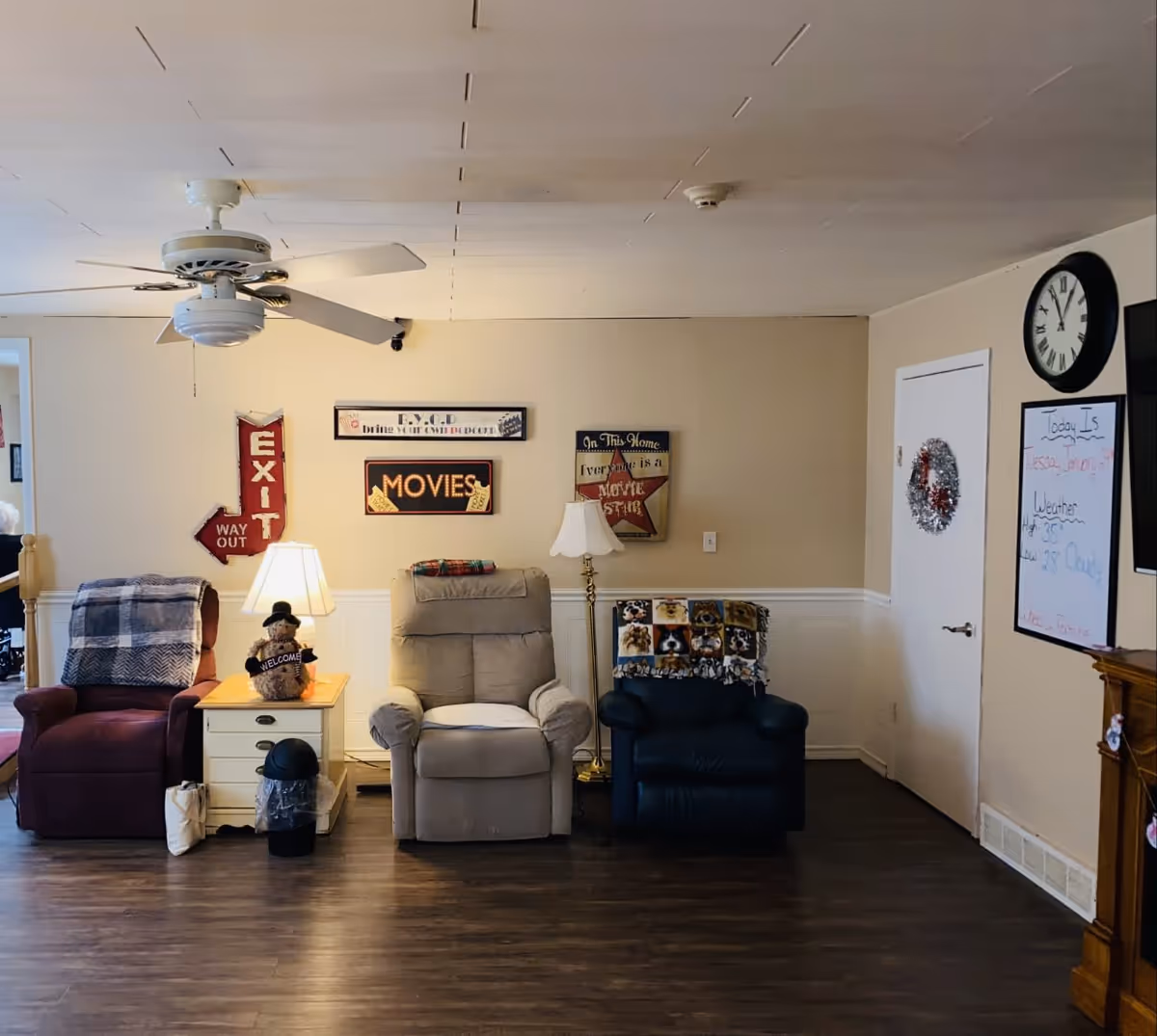 A cozy living room area with three recliner chairs in different colors (red, beige, and dark blue) arranged against a beige wall. There is a white side table with a lamp and a teddy bear holding a welcome sign between the red and beige chairs. The wall has decorative signs including an 'EXIT WAY OUT' arrow, a 'MOVIES' sign, and a sign that reads 'In This Home Everyone is a Movie Star.' A ceiling fan is visible above, and a clock and whiteboard with weather and date information are mounted on the right wall. The floor is dark wood, and there is a white door decorated with a wreath.