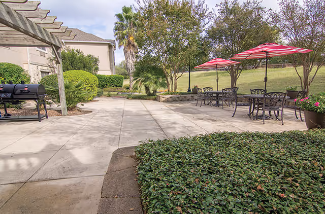Outdoor patio area at a senior living facility with metal tables and chairs under red and white striped umbrellas, a barbecue grill, greenery, trees, and a pergola structure.