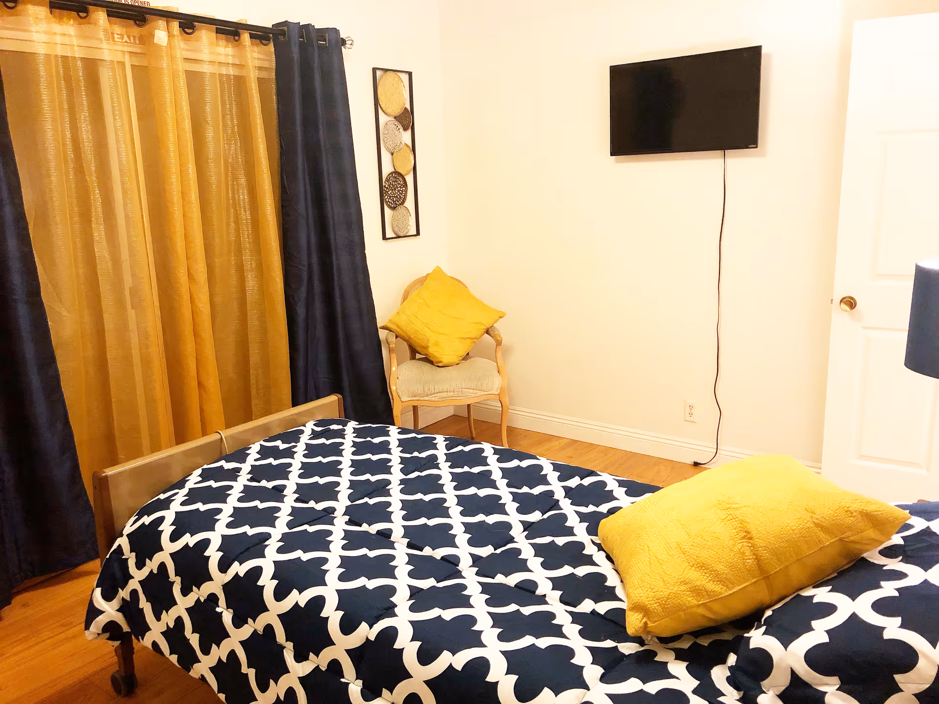 Cozy bedroom with a navy-and-white patterned bedspread, yellow pillows, a wall-mounted TV, curtains, and a chair.