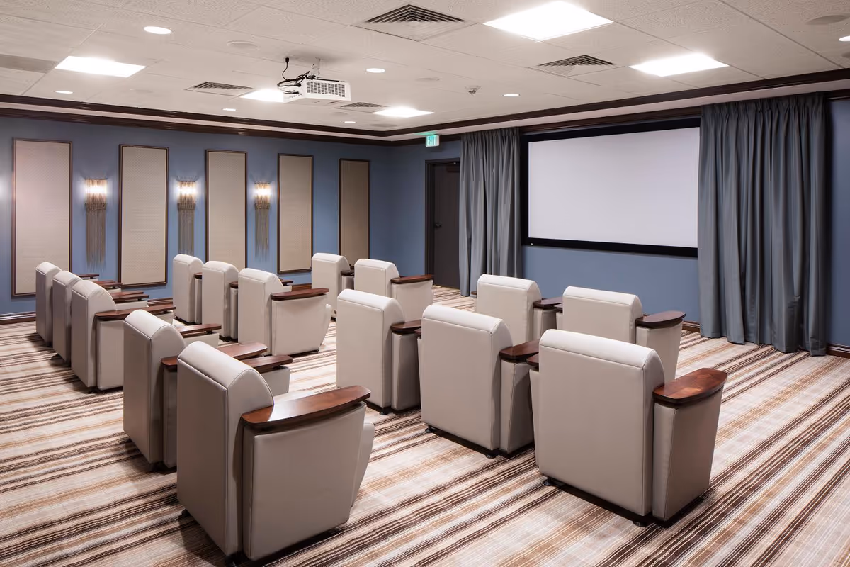 A small theater room with three rows of beige leather recliner chairs facing a large white projection screen. The walls are painted blue with decorative panels and wall sconces. The floor is carpeted with a striped pattern, and there is a ceiling-mounted projector above the seating area.