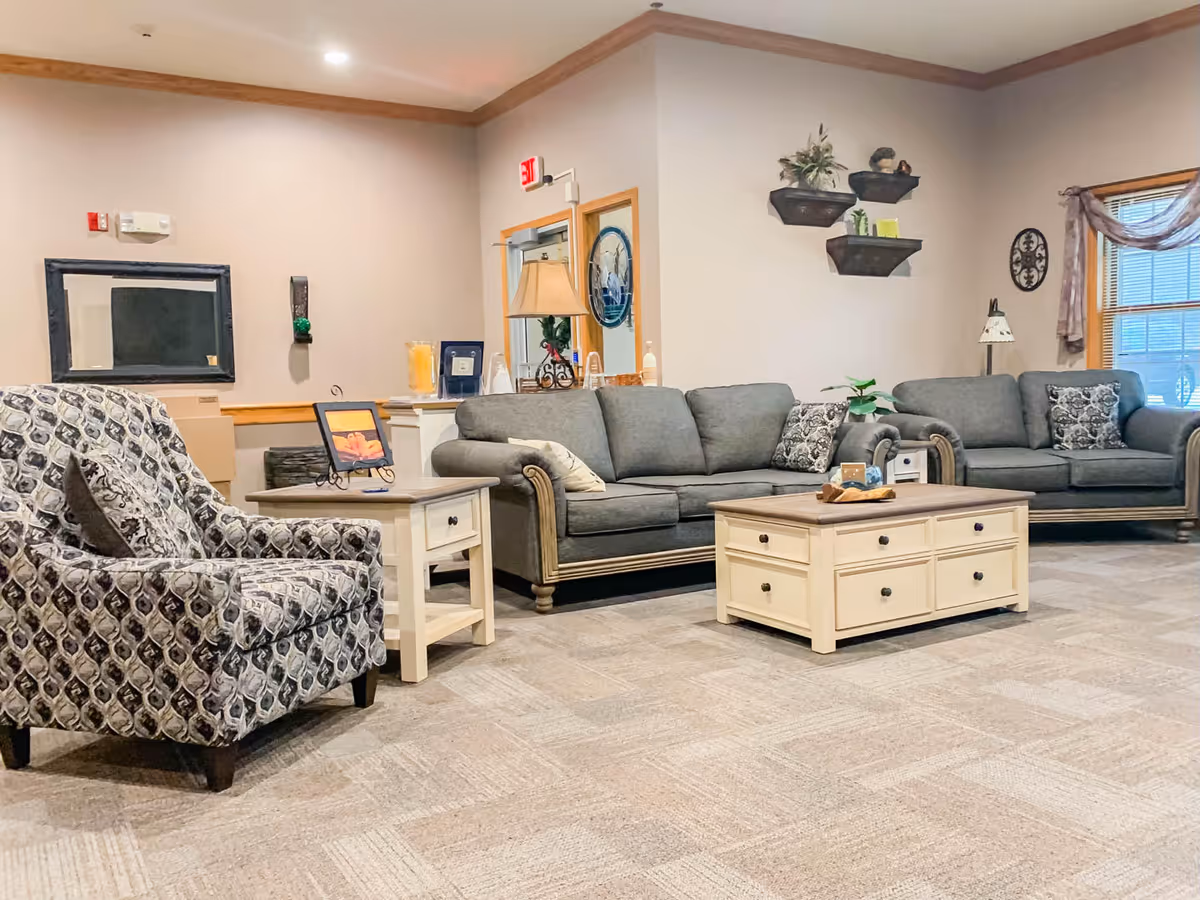 A cozy living room area in Meadow View Assisted Living featuring two gray sofas with patterned throw pillows, a patterned armchair, a wooden coffee table with drawers, and a side table with a framed picture. The room has beige walls, carpeted flooring, decorative wall shelves with plants, a window with curtains, and soft lighting.