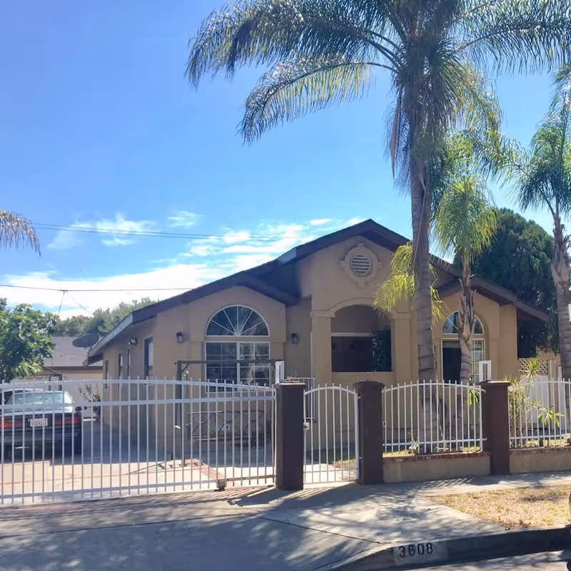 Exterior view of a single-story residential building with beige walls and a brown roof, surrounded by a white metal fence with brown pillars. There are palm trees in front of the building and a car parked in the driveway. The sky is clear and blue.