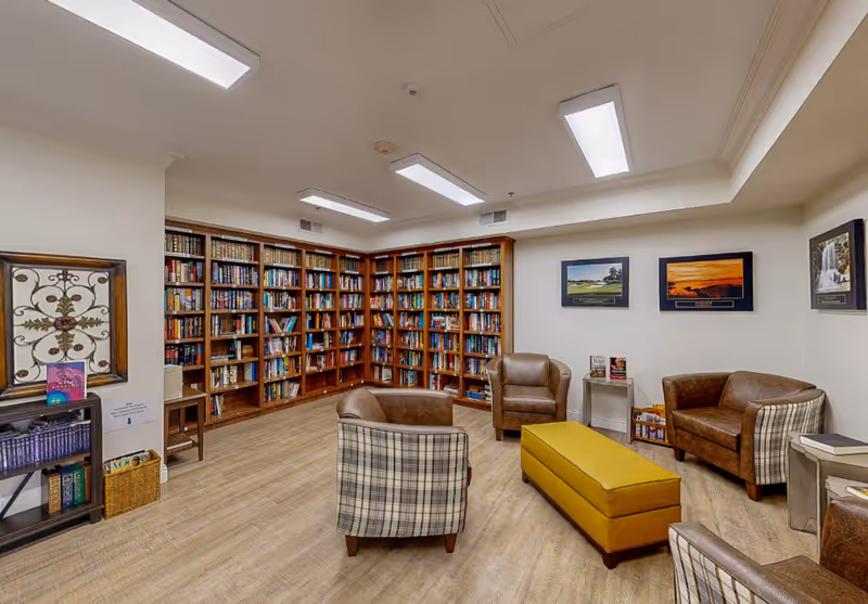 Cozy community library with bookshelves lining the walls, several armchairs and a yellow ottoman on wood floors.