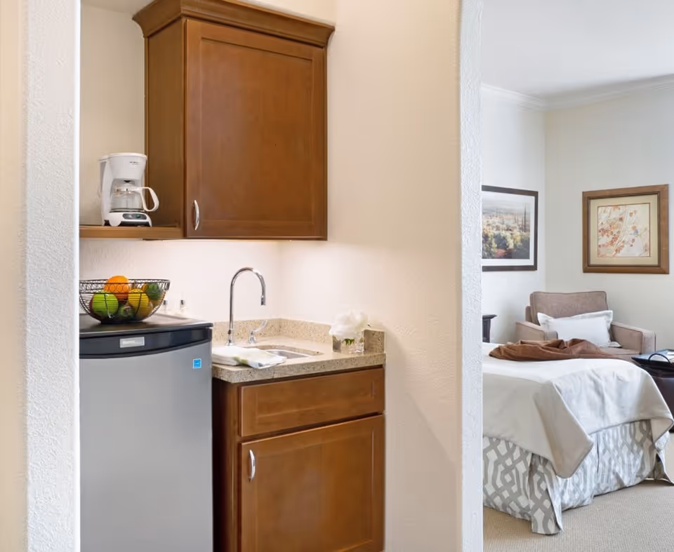 A small kitchenette area with wooden cabinets, a mini refrigerator, a coffee maker, a sink, and a bowl of fruit. In the background, there is a bedroom with a bed, an armchair, and framed artwork on the walls.
